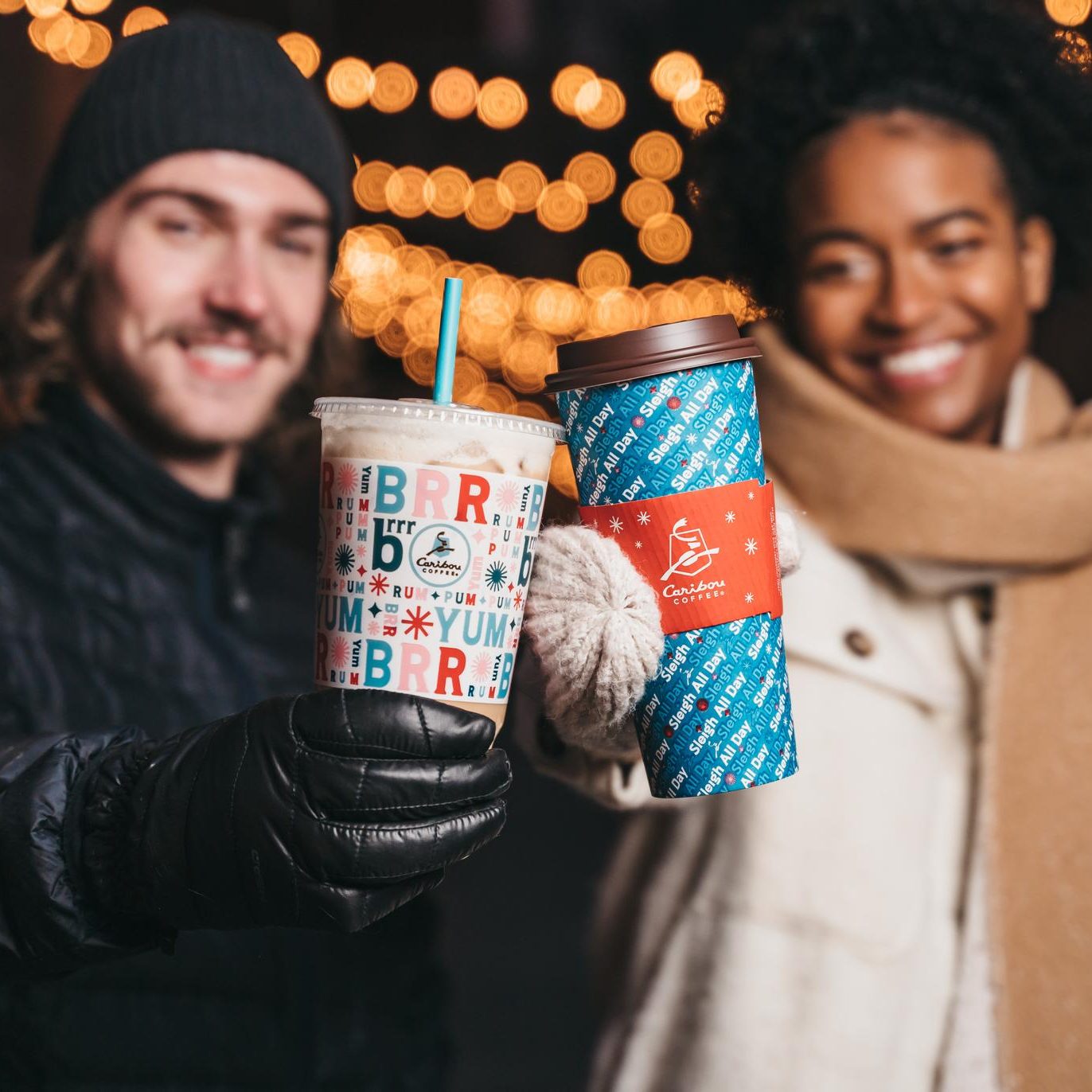 Two people holding Caribou Coffee Holiday cups, one cold cup, one hot. The cups say "Brrrum Pum Pum Yum" and "Sleigh All Day" as part of a pattern.
